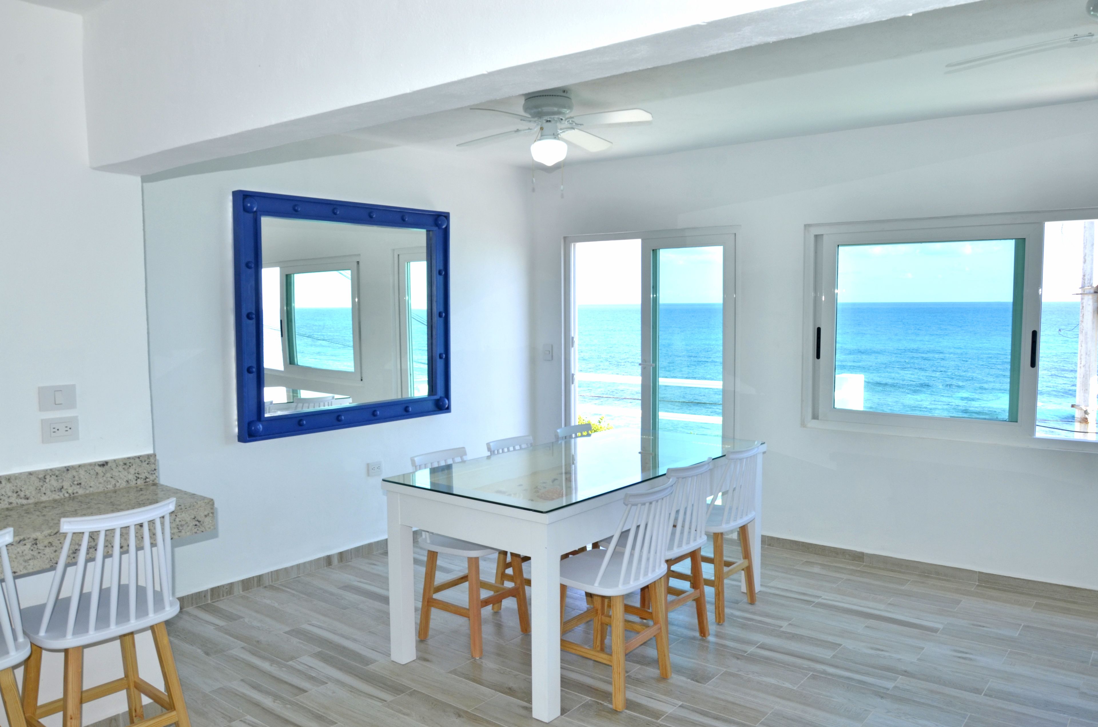 A spacious room with a glass table and white chairs, large windows with ocean views, and a ceiling fan.