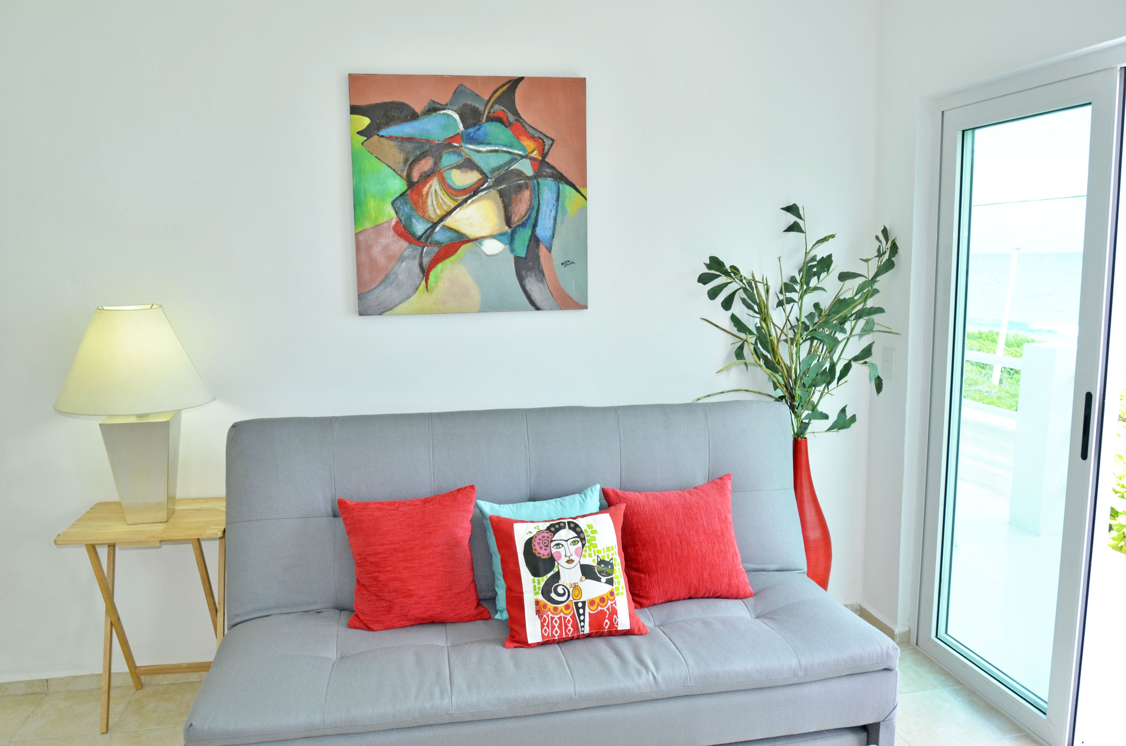 Um sofá cinzento com almofadas vermelhas e estampadas, uma luminária moderna e um quadro abstrato grande na parede.