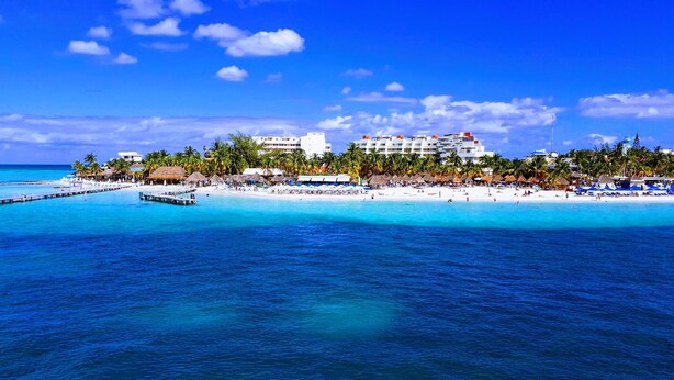 Un resort en la playa con un muelle que se adentra en aguas turquesas, playas de arena blanca y palmeras.