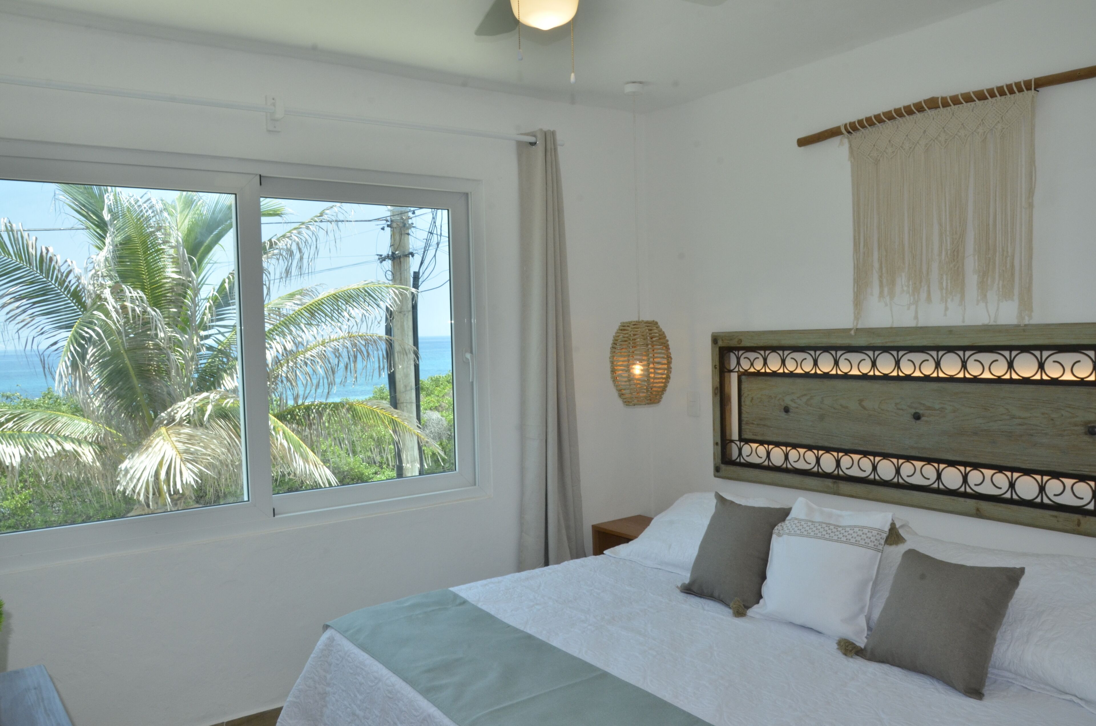 A bedroom with a large window, a bed with white and grey bedding, a wooden headboard, and a wall-mounted light fixture.
