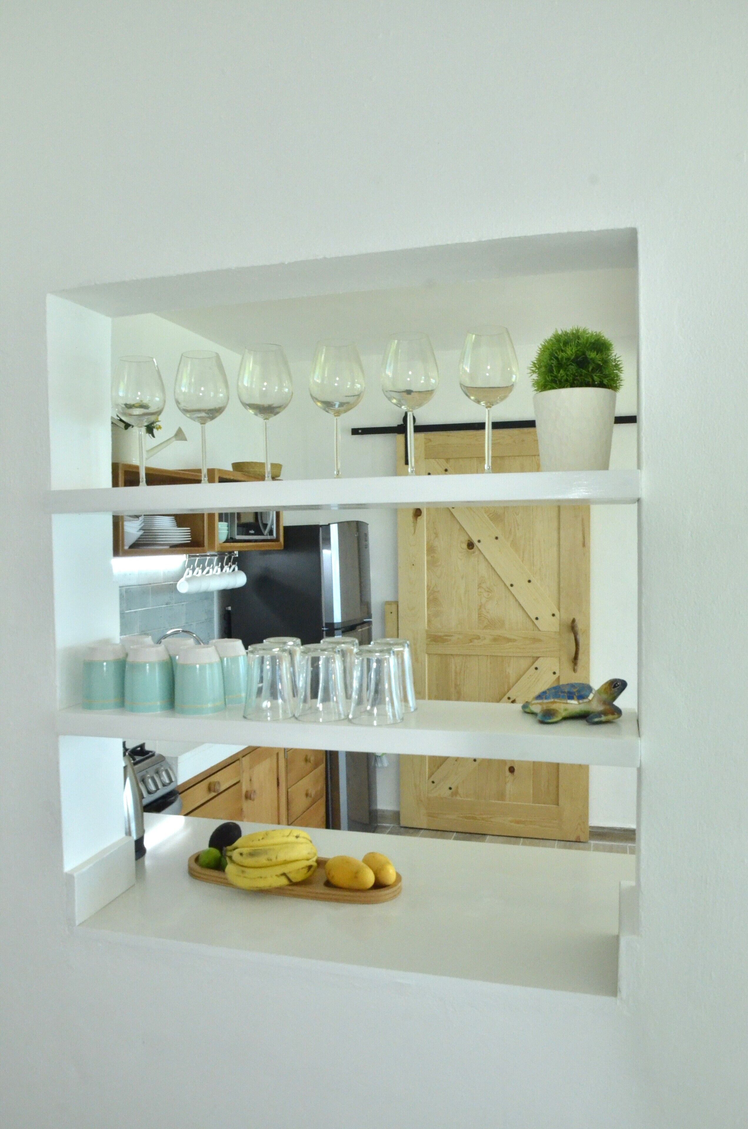 A kitchen with a wooden door, a fruit bowl with bananas, and a shelf with wine glasses and a potted plant.
