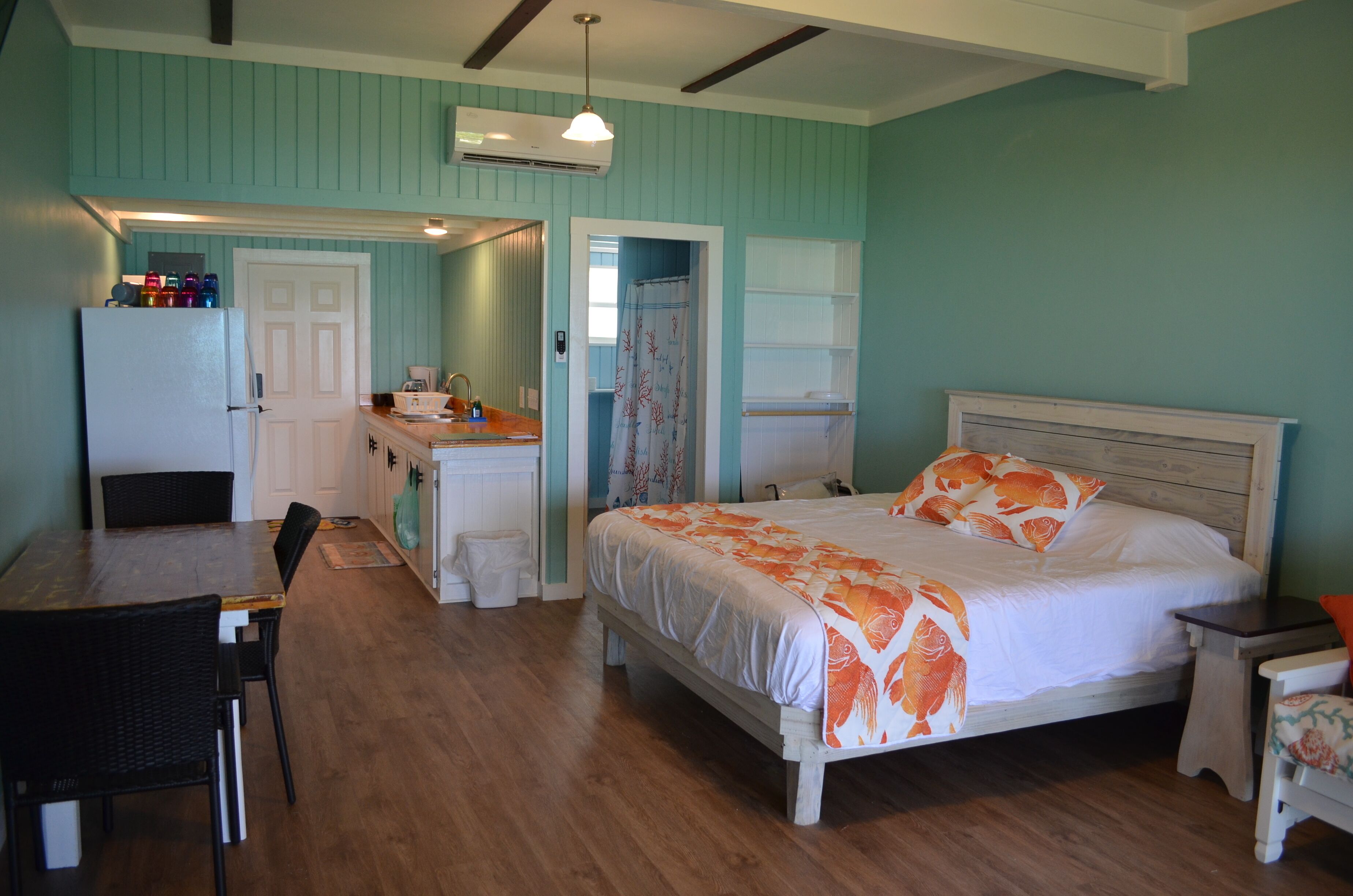 Un dormitorio con cama, mesita de noche, silla, pequeña mesa, escritorio, lavamanos, espejo y puerta.