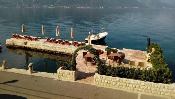 A waterfront patio with lounge chairs and umbrellas, a boat docked nearby, and mountains in the background. - Studio apartment for couples (Kotor)