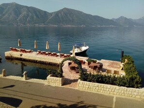 Un patio frente al agua con sillones de descanso y sombrillas, un barco amarrado cerca y montañas al fondo.