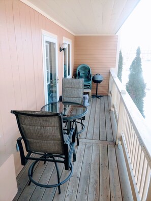 A wooden deck with a glass-top table and chairs, a small round table, and a chair with a backrest.