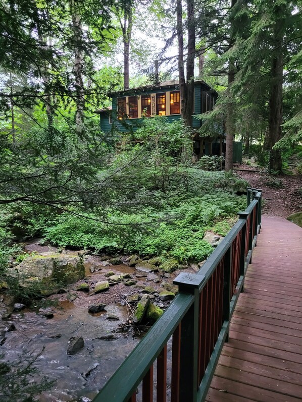 A wooden bridge leading to a cabin in a forested area with a stream.