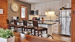 A modern kitchen with white cabinets, stainless steel appliances, a wooden dining table with chairs, and a wall clock.