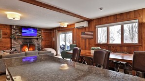 A cozy cabin interior with a stone fireplace, a television mounted above it, and a dining area with wooden tables and chairs.