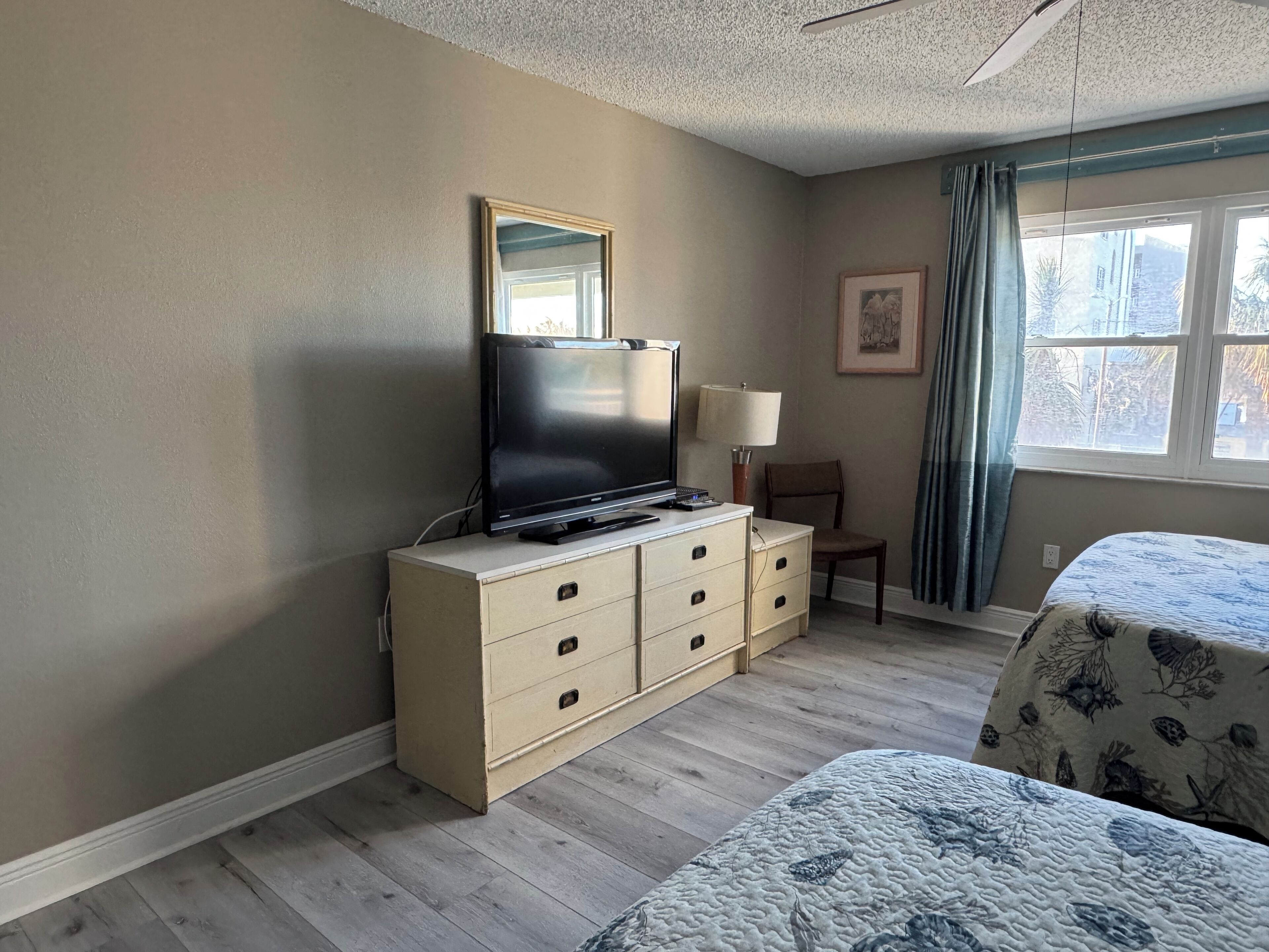 Quarto de hotel com cama, cômoda, televisão, poltrona e janela com cortinas.