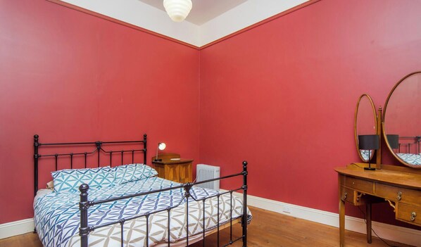 A bedroom with a red wall, a wooden dresser, a bed with a patterned bedspread, a nightstand with a lamp, and a mirror. - Central - Port Elliot
Historic Villa (Port Elliot)