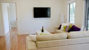 Un salon moderne avec un canapé beige, un téléviseur à écran plat fixé au mur et une petite table basse blanche sur laquelle se trouvent une bouteille et un verre.
