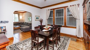 A dining area with a wooden table, chairs, and a rug. There is a window with curtains and a ceiling fan.