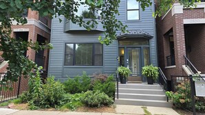 A two-story townhouse with a dark gray exterior, a front entrance with a glass door, and a small front yard with various plants.