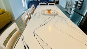 A modern kitchen with a large island, a dining area with a white marble table, and a cozy living space with a sofa and TV.