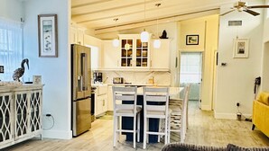A modern kitchen with white cabinets, a central island with chairs, a ceiling fan, and a dining area with a yellow sofa.