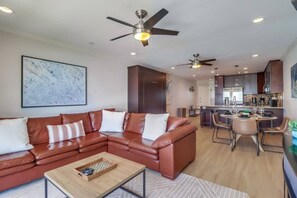 A modern living room with a sectional sofa, a coffee table, and a dining area with a bar. There are ceiling fans and recessed lighting.