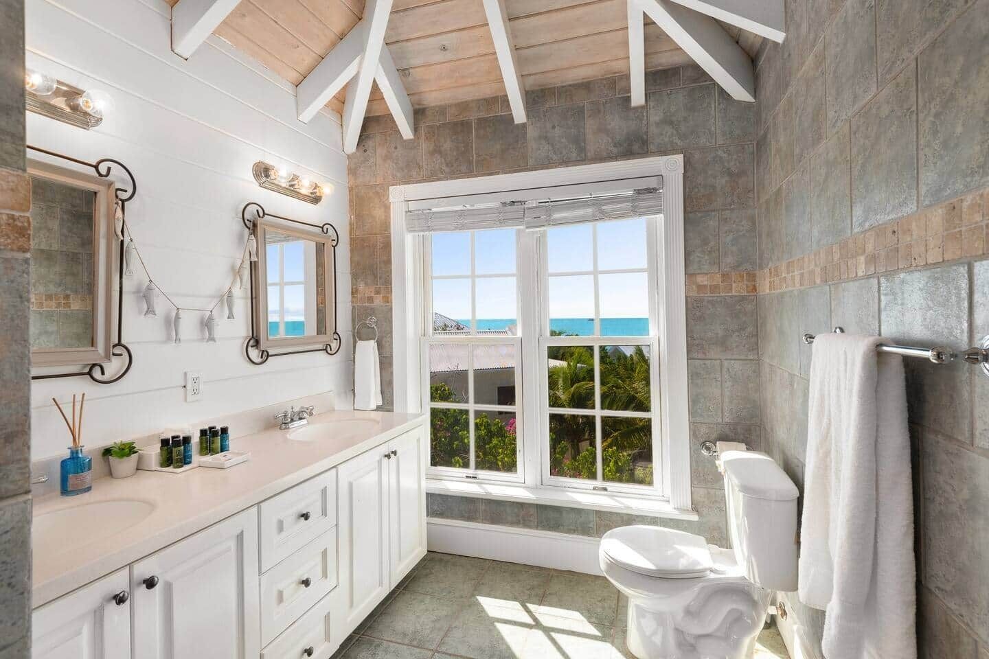 A bathroom with a large window, white fixtures, and a view of the ocean.