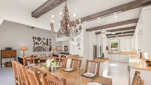 A spacious dining area with a wooden table set for six, surrounded by wooden chairs. A chandelier hangs from the ceiling, and the room features exposed wooden beams.