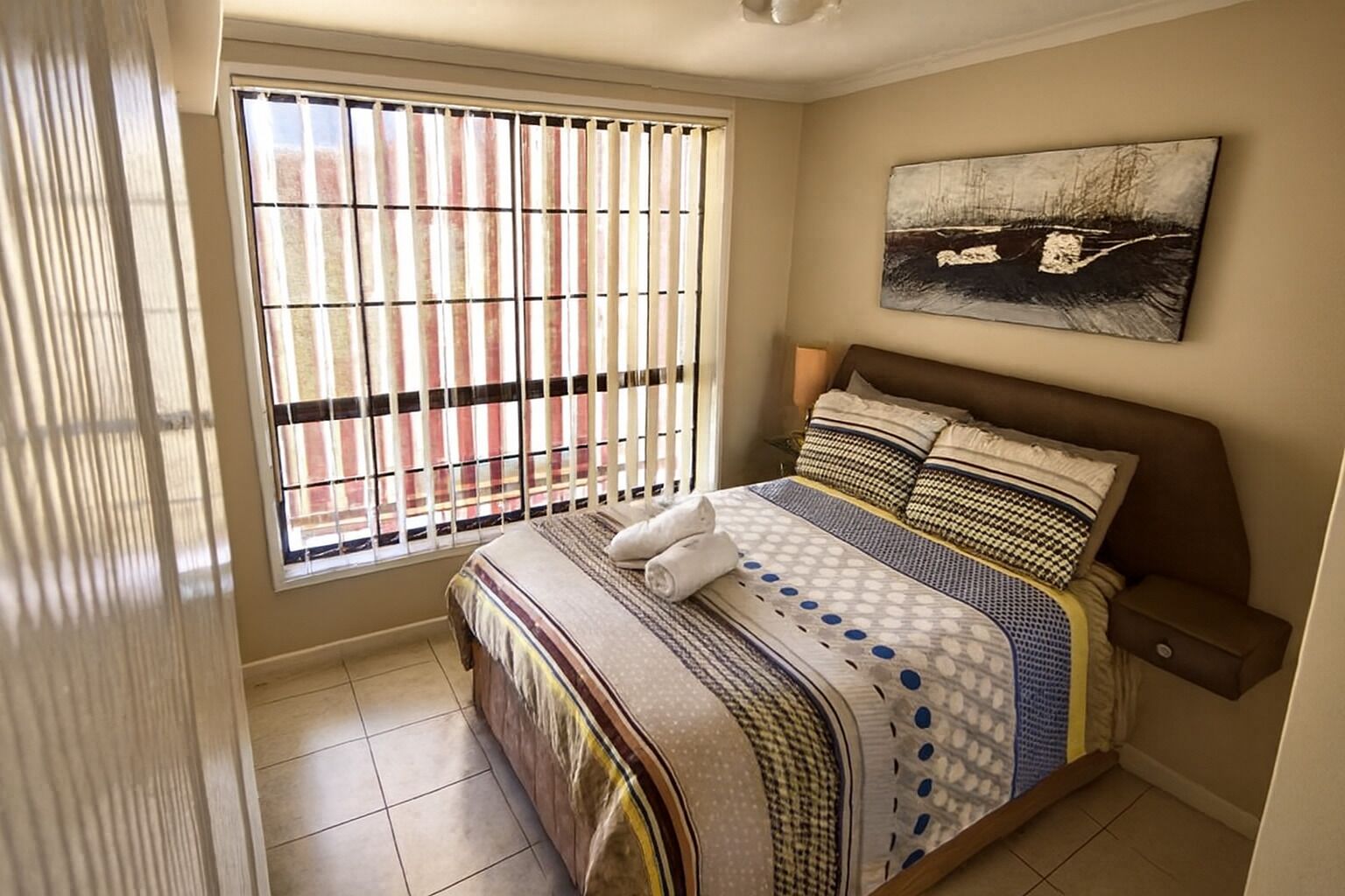A bedroom with a large bed, a window with blinds, a painting on the wall, and a tiled floor.