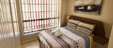A bedroom with a large bed, a window with blinds, a painting on the wall, and a tiled floor.