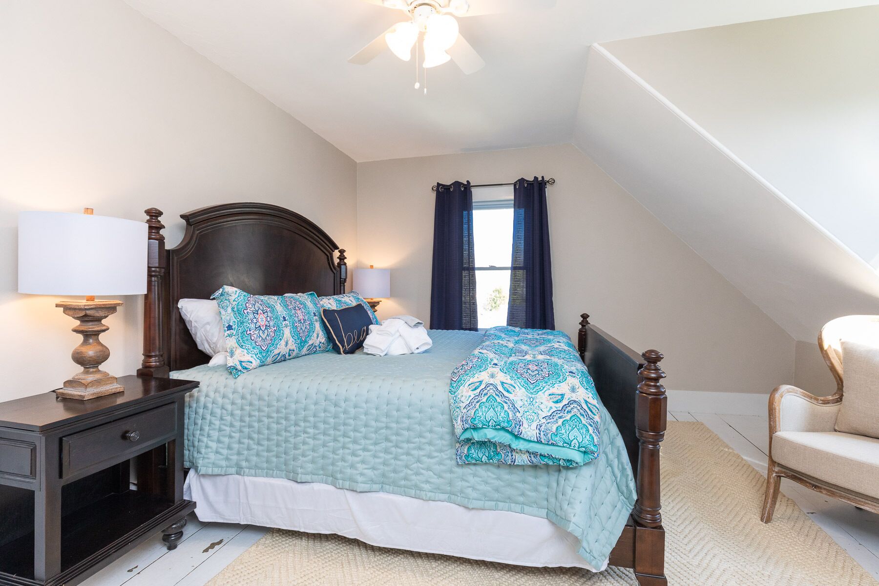 A bedroom with a bed, nightstands, a chair, a window with curtains, and a ceiling fan.