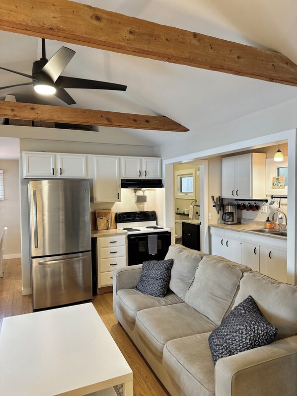Una cocina moderna con armarios blancos, un refrigerador de acero inoxidable y una estufa negra. En la zona de estar, hay un sofá color beige con almohadones estampados.