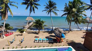 A pool area with clear blue water, surrounded by lounge chairs and palm trees, overlooking a beach with a thatched-roof hut. - Luxury, Beachfront, Home with Resort-amenities, Housekeeping & FREE GOLF CART. (Hopkins)