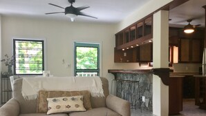 Un salon avec un canapé beige, un plaid blanc et un coussin décoratif. On y trouve également un coin cuisine avec des meubles en bois et un pare-feu en pierre.