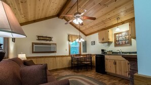 A cozy living room with a sofa, a dining table, and chairs, a kitchen with wooden cabinets and a window, and a ceiling fan.