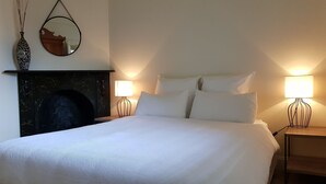 Ein ordentlich bezogenes Bett mit weißen Leinen, zwei Nachttischlampen, ein an der Wand befestigter Spiegel und eine dekorative Vase.