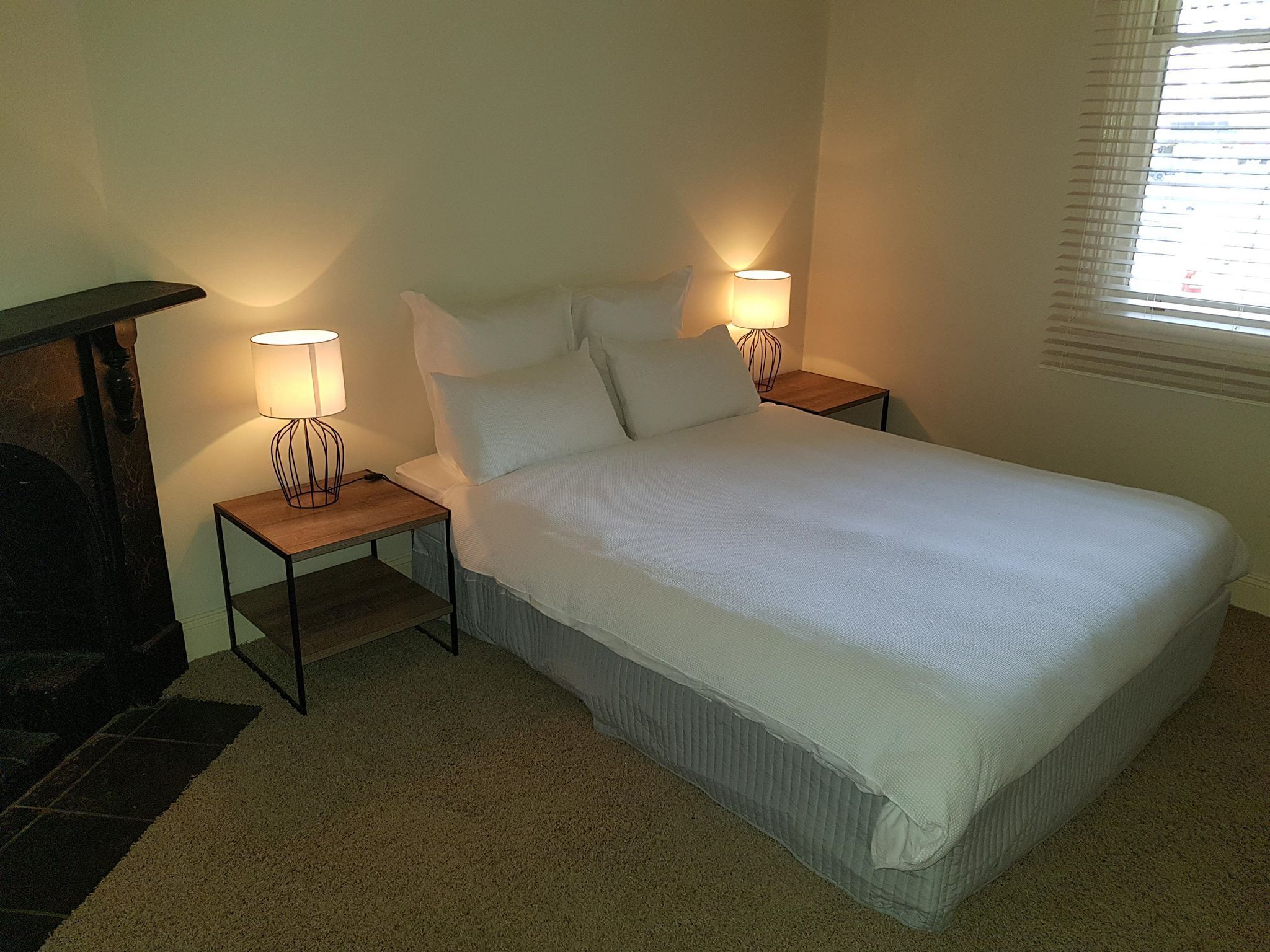 A bedroom with a bed, two bedside tables with lamps, a carpeted floor, and a window with blinds.