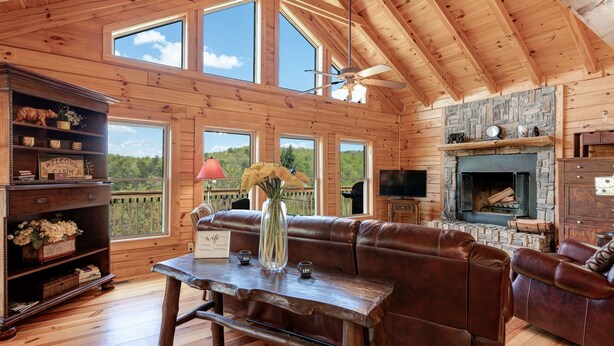 Interior de una cabaña con techo de madera, ventanas amplias, chimenea, sofá de cuero y una mesa con un florero y una lámpara.