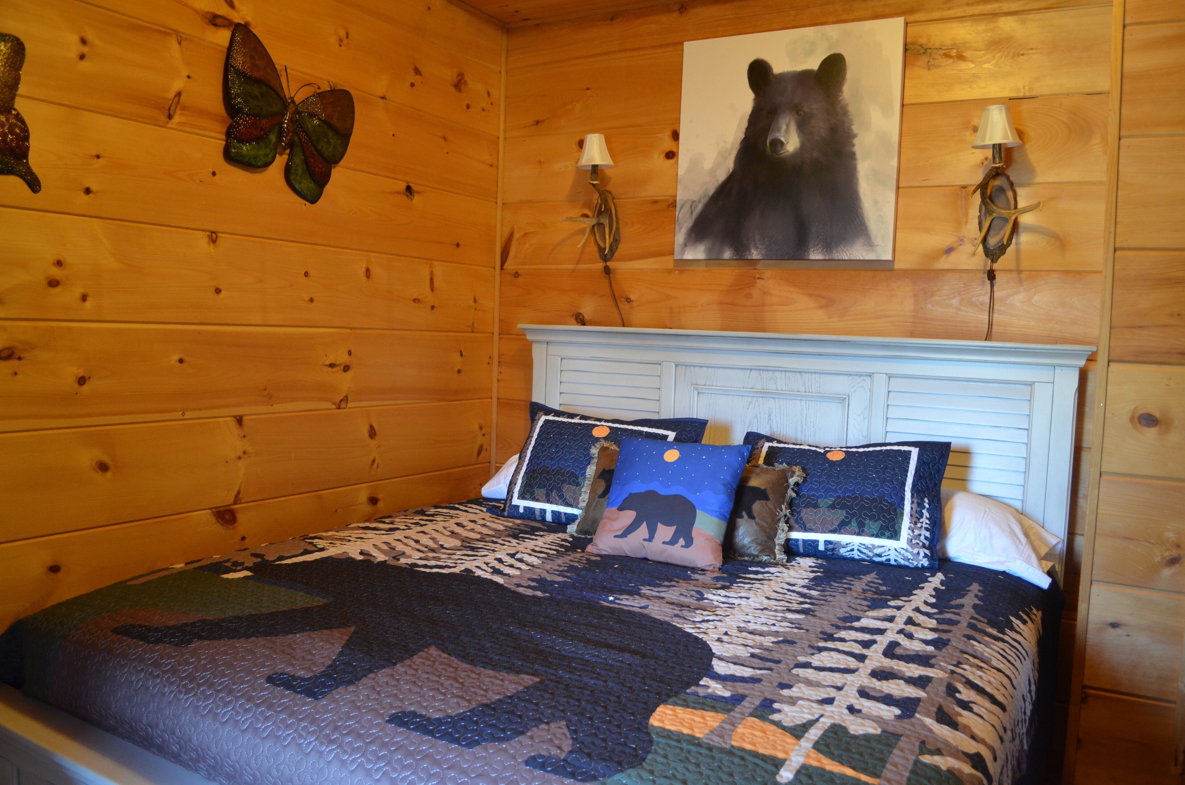 A wooden cabin bedroom with a bed, a bear-themed pillow, a bear painting on the wall, and a butterfly decoration.