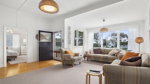 A spacious living room with a dark wooden door, two armchairs, a sofa, and a coffee table. Large windows allow natural light and offer views of the outdoors. - Wisteria Cottage - opposite beach (Indented Head)