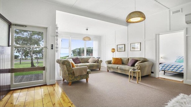 A spacious living room with a wooden floor, two sofas, a small side table, and a large window offering a view of the outdoors. - Wisteria Cottage - opposite beach (Indented Head)
