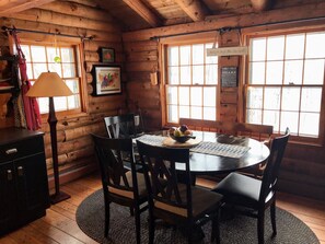 A cozy cabin interior with a wooden dining table, chairs, a lamp, and a window with a view.
