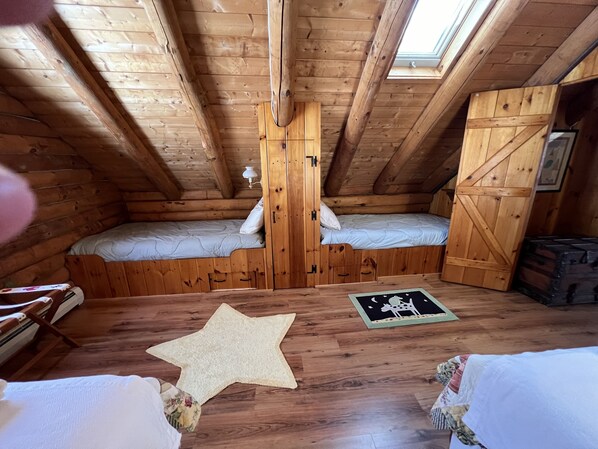 A cozy attic bedroom with wooden walls and a sloped ceiling. There are two beds with white bedding, a star-shaped rug, and a cow-patterned rug on the floor.