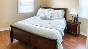 Une chambre à coucher avec un lit en bois, un ventilateur de plafond, une table de chevet avec une lampe et deux fenêtres équipées de stores.