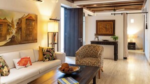 Una sala de estar con un sofá, una mesa de centro con un libro y un cuenco, un sillón estampado, un escritorio con una lámpara y un cuadro en la pared.