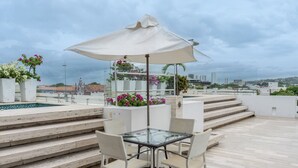 Una terraza en la azotea con una mesa y sillas de vidrio, rodeada de plantas en macetas y una piscina.