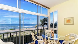 Un balcon avec une table en verre et des chaises en osier, donnant sur une plage et l’océan.