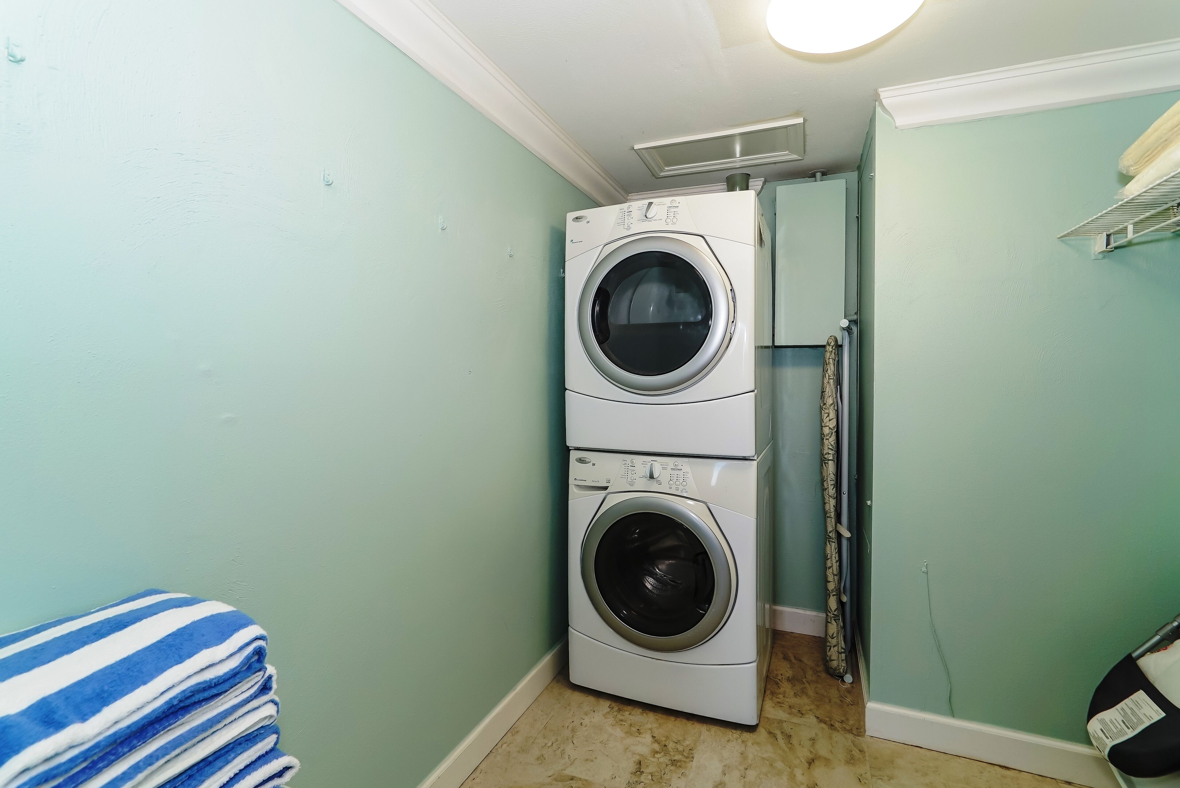 A laundry room with two stacked washing machines, a blue-striped towel, and a door leading to another room.