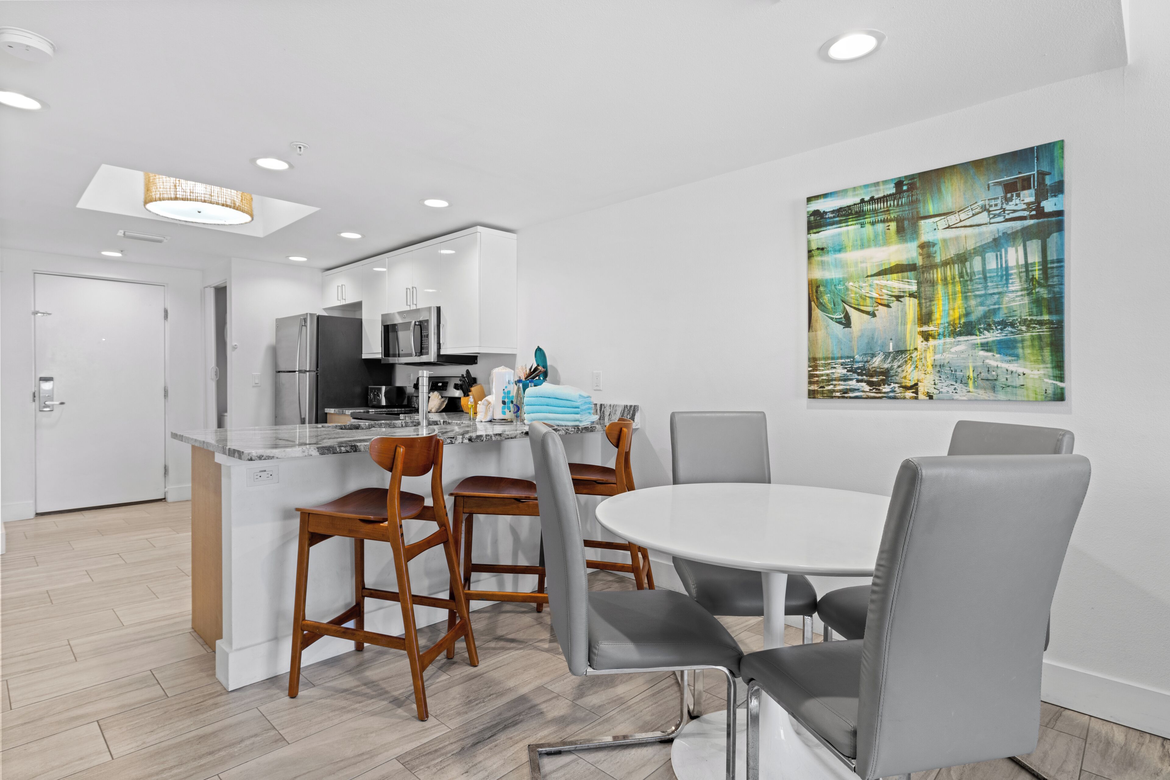 A modern kitchen with white cabinets, a stainless steel refrigerator, and a built-in microwave. There is a dining area with a white table and grey chairs.