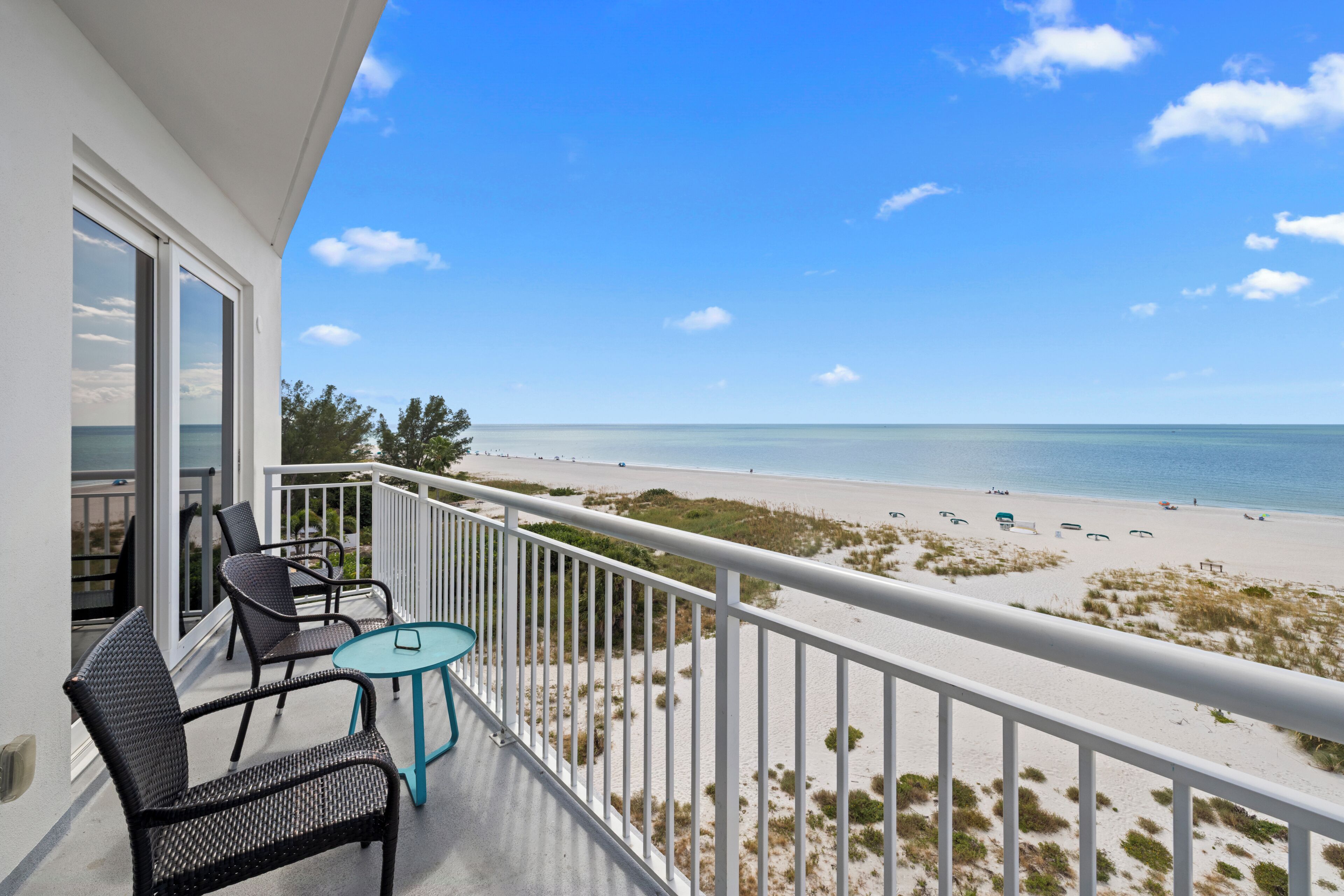 A balcony with wicker chairs and a small table, overlooking a beach with sand dunes and the ocean.