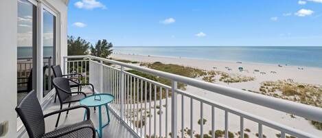 A balcony with wicker chairs and a small table, overlooking a beach with sand dunes and the ocean.