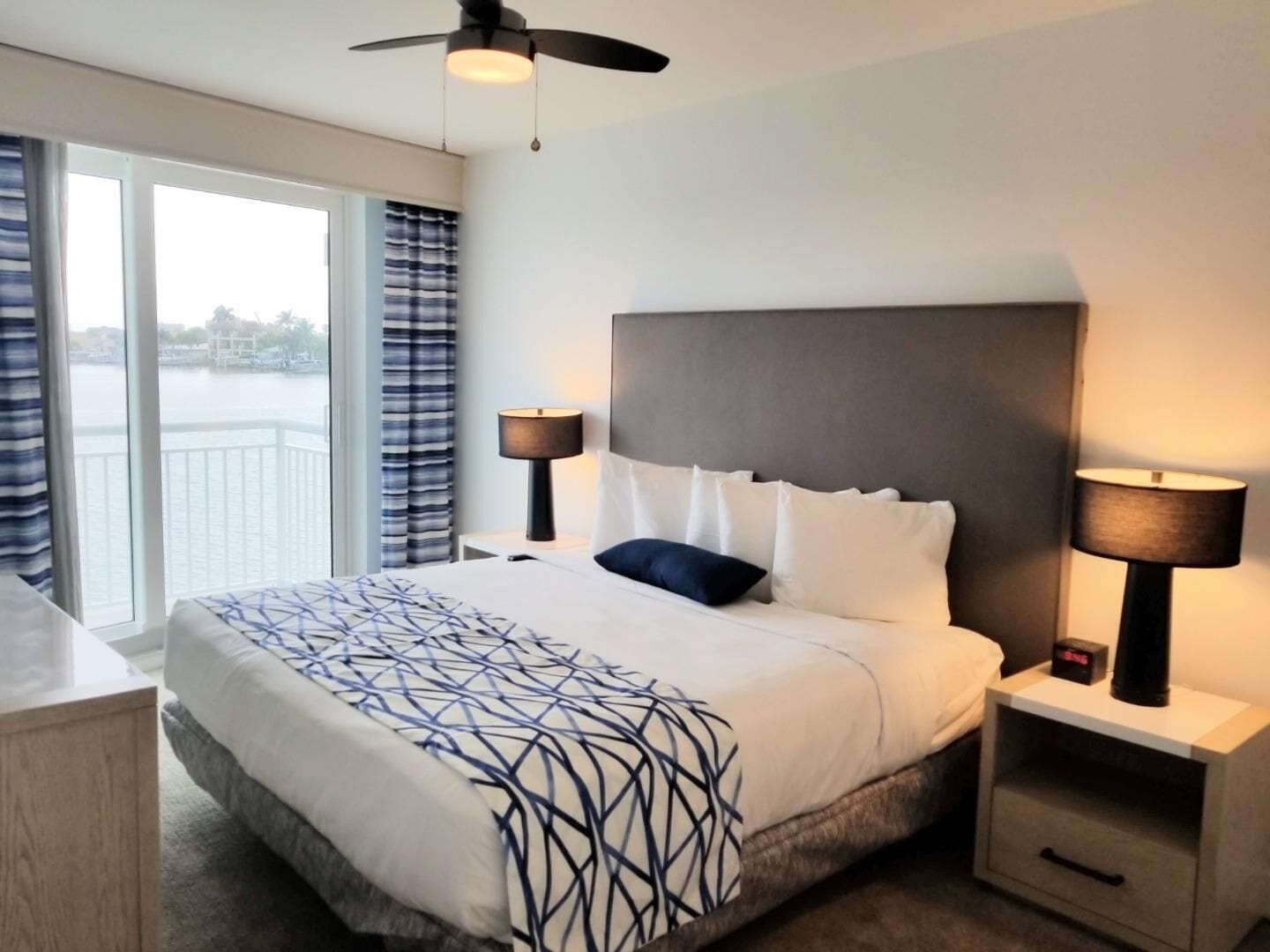 Een hotelkamer met een groot bed, twee nachtkastjes met lampen, een plafondventilator en uitzicht op een wateroppervlak door grote ramen.