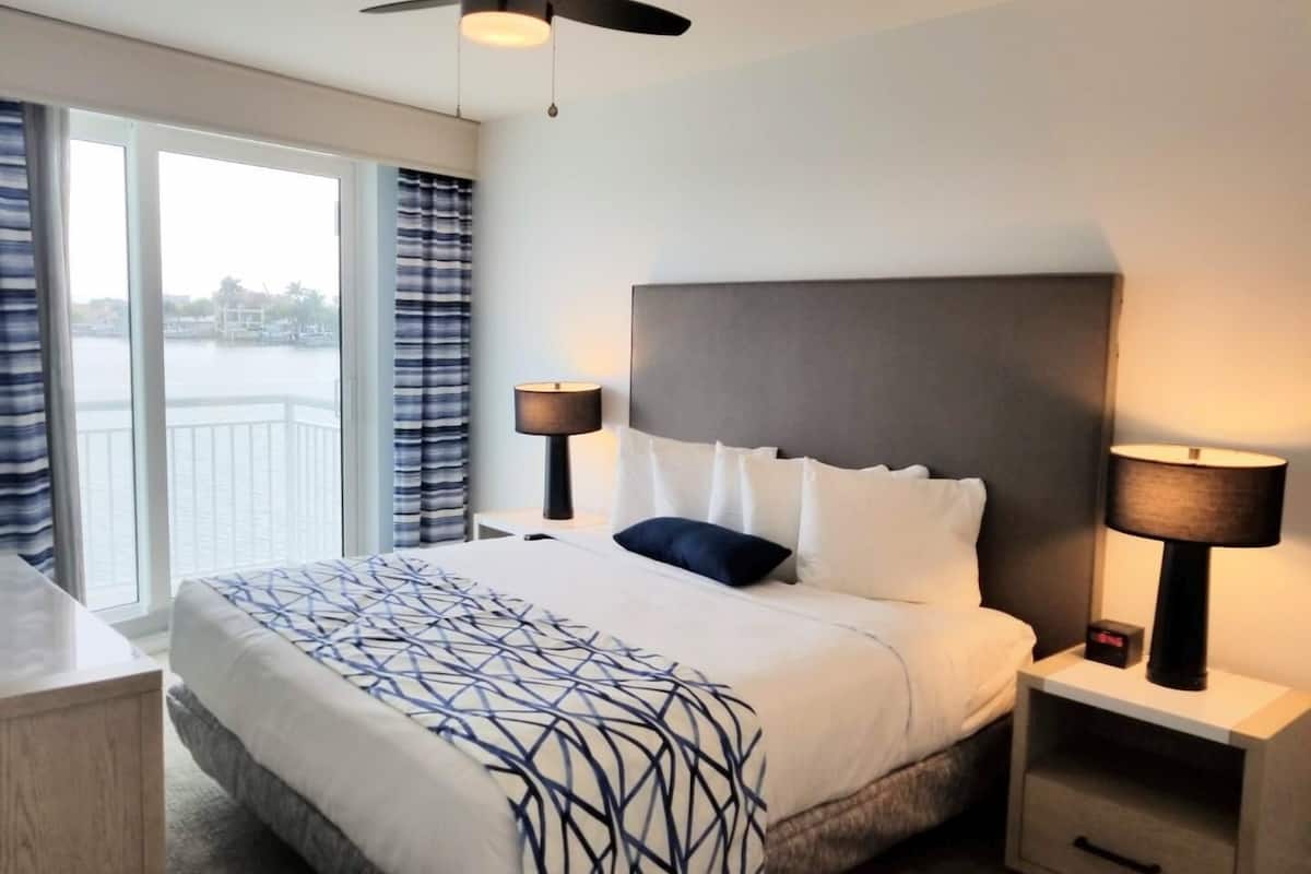Habitación de hotel con una cama grande, dos mesitas de noche con lámparas, un ventilador de techo y vistas a una masa de agua a través de grandes ventanales.