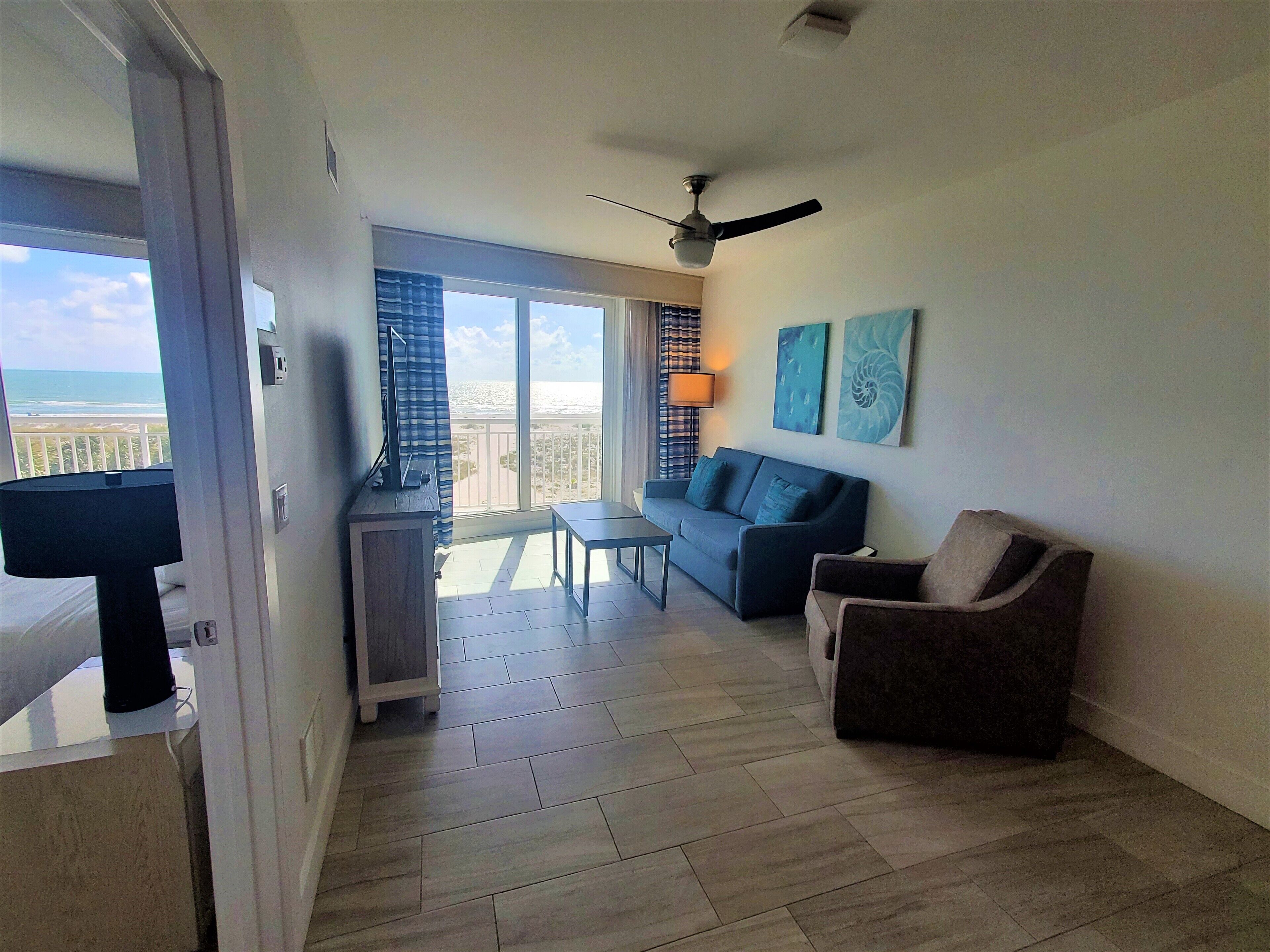 A living room with a sofa, armchair, and a small table. There is a balcony with a view of the ocean.