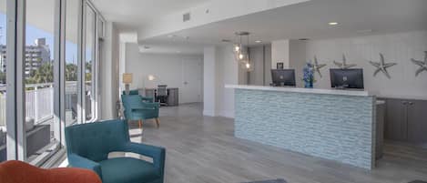 A modern hotel lobby with a reception desk, teal armchairs, and a large window offering a city view.