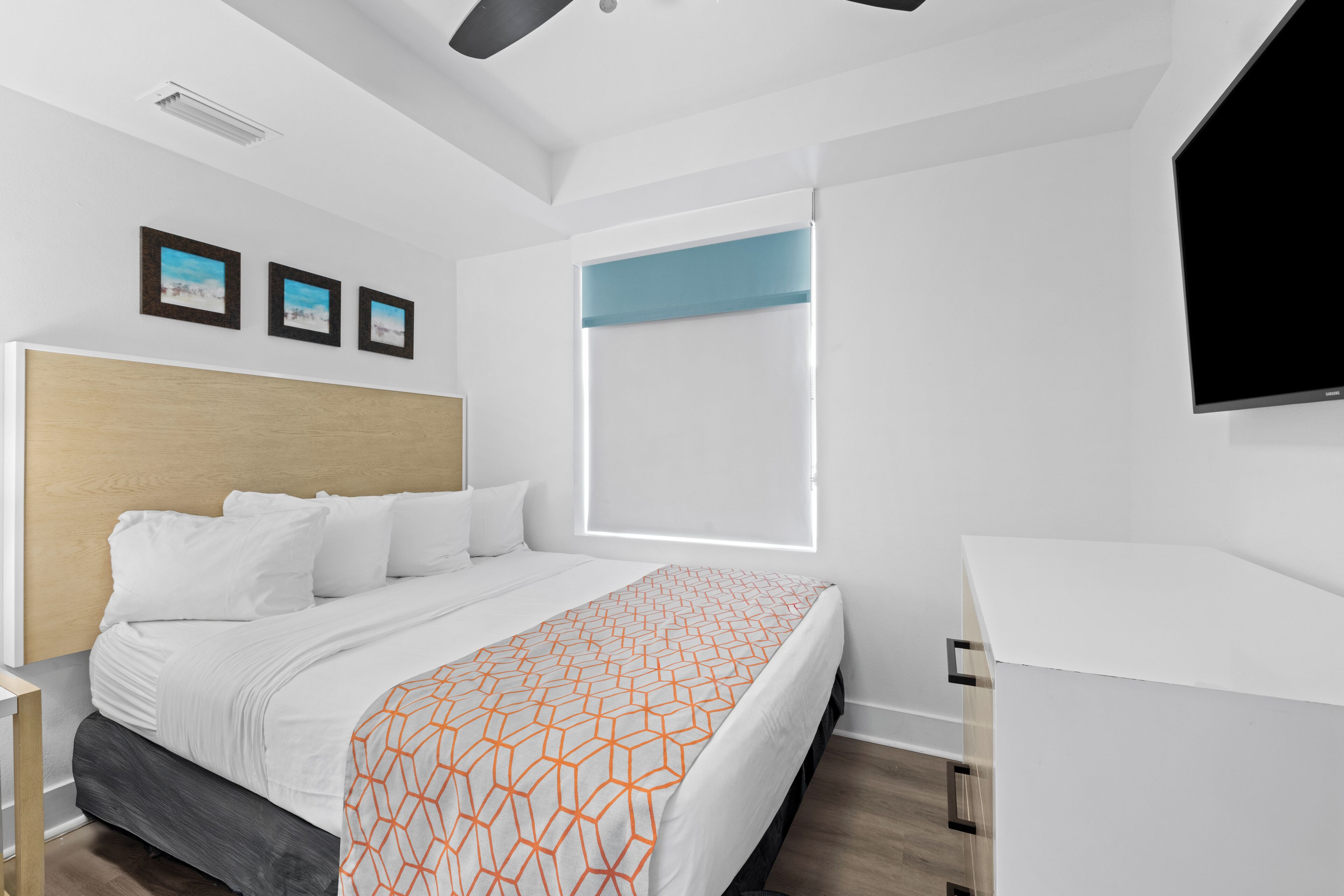 A modern hotel room with a wooden headboard, a large bed with white bedding, a desk, and a flat-screen TV mounted on the wall.